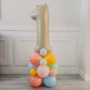 May include: A large gold number one balloon sits atop a cluster of colorful balloons. The balloons are in shades of peach, blue, yellow, and green, creating a festive display. This arrangement is suitable for a first birthday celebration.