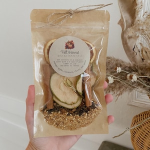Peut inclure: Un sachet transparent nou&eacute; avec de la ficelle, contenant un kit de pot-pourri pour cuisini&egrave;re Fall Harvest. Le kit comprend des tranches d'orange s&eacute;ch&eacute;es, des tranches de pomme, des b&acirc;tons de cannelle, des clous de girofle et de l'anis &eacute;toil&eacute;. Le sachet a une &eacute;tiquette circulaire avec du texte.