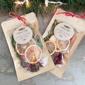 Puede incluir: Dos bolsas de papel marr&oacute;n con ventanas de pl&aacute;stico transparente que contienen rodajas de naranja secas, ar&aacute;ndanos rojos, palitos de canela, an&iacute;s estrellado y ramitas de romero. Las bolsas est&aacute;n atadas con hilo rojo y marr&oacute;n y tienen etiquetas circulares blancas con el texto "Christmas Magic Holiday Simmer Kit" y "Holiday Magic Simmer Kit" impreso en ellas.