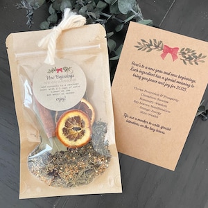 May include: A brown paper bag with a clear window, containing dried orange slices, cinnamon sticks, bay leaves, and other herbs. The bag is tied with twine and has a label that reads "New Beginnings New Year Simmer Pot". A separate card with instructions and a list of ingredients and their meanings is included.