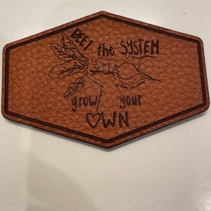 May include: A brown leather patch with a hexagonal shape and a dark brown border. The patch features the words "BEET the SYSTEM grow your OWN" with a beet and leaf illustration.