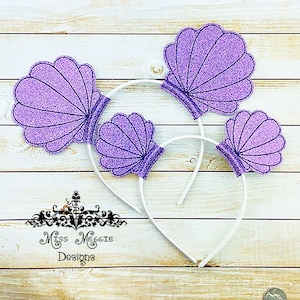 Op de afbeelding: Drie paarse glitter schelp haarbanden op een witte houten achtergrond. De haarbanden zijn wit en de schelpen zijn paars met een glinsterende afwerking.