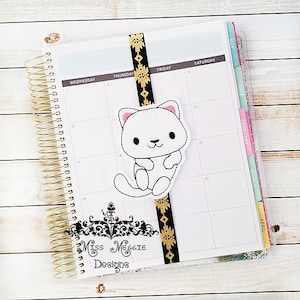May include: A white cat with pink ears and a black nose is embroidered on a black and gold bookmark. The bookmark is attached to a planner with a gold coil binding. The planner has a white cover with a monthly calendar printed on it. The text "Miss MEGGIE Designs" is printed on the planner.