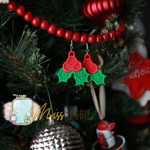 May include: A pair of green and red holly berry earrings. The earrings are made of felt and are hanging from a Christmas tree.