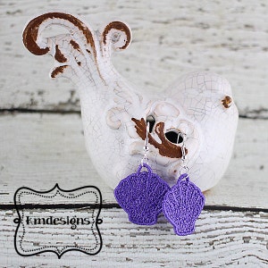 May include: Purple crocheted seashell earrings hanging from a white bird figurine on a white wooden surface.