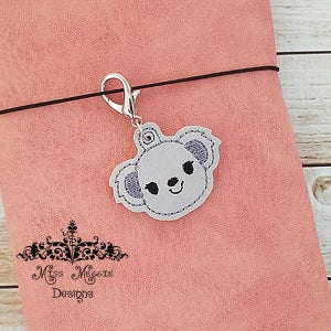 May include: A silver charm with a gray embroidered koala face. The charm is attached to a black elastic cord and is hanging from a pink leather notebook.