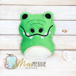 May include: Green plush alligator head pillow with embroidered eyes, teeth, and nostrils. The pillow is on a white wood background.
