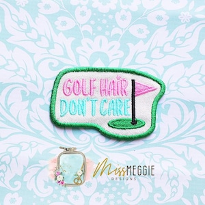 May include: Embroidered patch with the text "GOLF HAIR DON'T CARE" in pink and teal. The patch features a golf flag and green border. The background is a light blue floral pattern.