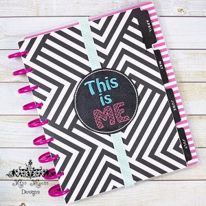 May include: A black and white planner with a chevron pattern and a pink and black "This is Me" circle sticker. The planner has a clear plastic cover and a pink and white ribbon bookmark. The planner has a set of dividers with the months April, May, June, and July.