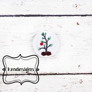 Puede incluir: Un círculo de fieltro blanco con un árbol de Navidad verde bordado con un adorno rojo. El árbol está sobre una base de fieltro marrón. El círculo tiene un borde blanco.