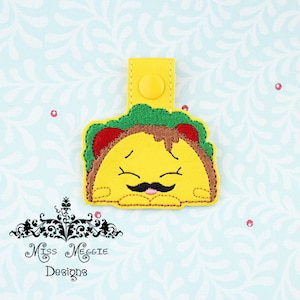May include: A yellow felt taco with a smiling face, brown mustache, and pink tongue. The taco has green lettuce, brown meat, and red tomato filling. The taco is attached to a yellow felt loop with a yellow button.