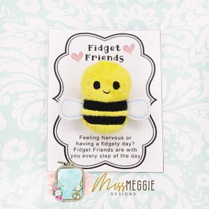 May include: A plush yellow and black bee with white wings is the focal point of this image. The bee is on a white card with the words "Fidget Friends" and a message about fidgety days. The card is decorated with pink hearts and a decorative border.