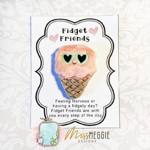 Könnte beinhalten: Ein rosa, plüschiger Eiswaffel-förmiger Zappelspielzeug mit herzförmiger Sonnenbrille, auf einer weißen Karte mit dem Text "Fidget Friends". Die Karte enthält auch den Text "Feeling Nervous or having a fidgety day? Fidget Friends are with you every step of the day."