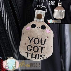 Peut inclure: Un porte-clés en peluche en forme de pomme de terre souriante avec le texte "YOU GOT THIS". Le porte-clés a des yeux noirs, des joues roses et un bouton marron sur la sangle. Une petite image montre le dos du porte-clés.