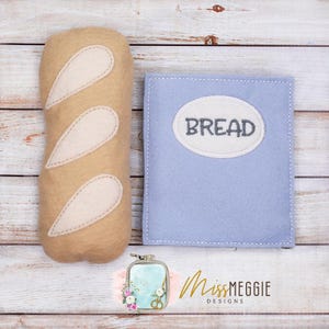 May include: A felt bread loaf with three white accents and a light blue bread storage bag. The bag has an oval patch with the word "BREAD" stitched in gray. The image is on a light wood surface.