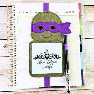 May include: A green felt bookmark with a purple felt Teenage Mutant Ninja Turtles mask, a white note pad with the text "Miss Maggie Designs" and a black pen.