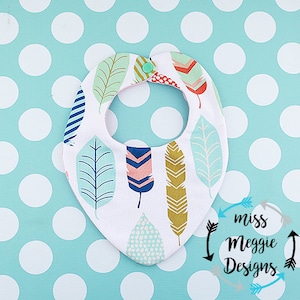 May include: A white baby bib featuring a feather design in blue, green, pink, and gold. The bib has a snap closure and is set against a light blue background with white polka dots. The bib is from Miss Meggie Designs.