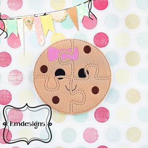 May include: A brown felt cookie puzzle with a pink bow, chocolate chips, and a smiling face. The cookie is on a white background with colorful polka dots.