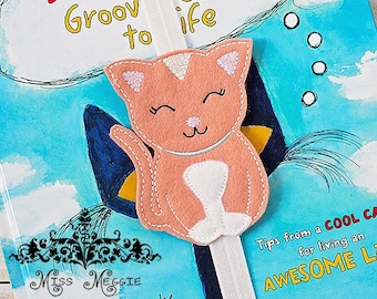 May include: A pink felt cat bookmark with white accents and embroidered eyes. The bookmark is attached to a white elastic band. The bookmark is on top of a book with the title "Pete's Guide to Life" and the text "Tips from a COOL CAT for living an AWESOME LIFE".
