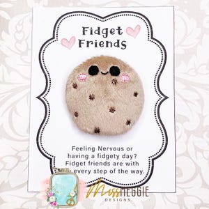 May include: A brown plush fidget toy shaped like a cookie with a smiling face and pink cheeks. The toy is on a white card with the text "Fidget Friends" and "Feeling Nervous or having a fidgety day? Fidget friends are with every step of the way."