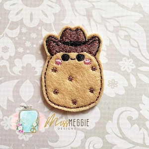 Puede incluir: Aplicación de fieltro beige con forma de personaje de dibujos animados con sombrero de vaquero marrón. El personaje tiene ojos negros, mejillas rosas y detalles bordados. La aplicación está cosida con hilo marrón.