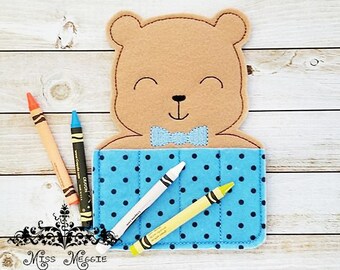May include: A felt crayon holder shaped like a brown bear with a blue bow tie. The holder has a blue polka dot fabric pocket with four crayons inside.