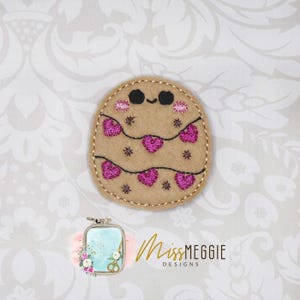 Peut inclure: Applique en feutre beige avec un visage souriant, des joues roses et une guirlande de cœurs roses. L'applique est brodée de fil noir et comporte de petits accents d'étoiles. Le design est circulaire et a une bordure cousue.
