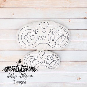 Puede incluir: Dos almohadas de tela blanca con forma de mandos de videojuegos. La almohada más grande tiene 12 pulgadas de ancho y la más pequeña 8 pulgadas de ancho. Ambas almohadas tienen una forma de corazón en la parte superior y un contorno negro simple de los botones del mando.