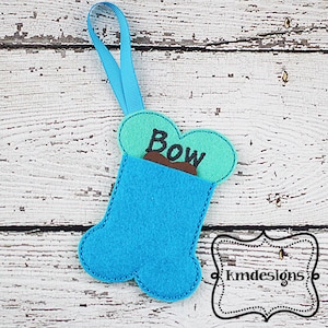 May include: A blue felt bone-shaped ornament with a green felt pocket on the front. The pocket has the word "Bow" embroidered on it. The ornament has a blue ribbon for hanging.