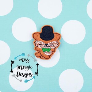 May include: Embroidered felt appliqué of a fox wearing a black top hat and a green bow tie. The fox is brown with white accents. The appliqué is on a white background with grey polka dots.