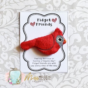May include: A plush red cardinal fidget toy with a gray and black eye, resting on a white card. The card reads "Fidget Friends" with hearts and a message about fidgety days. The MissMEGGIE DESIGNS logo is at the bottom.