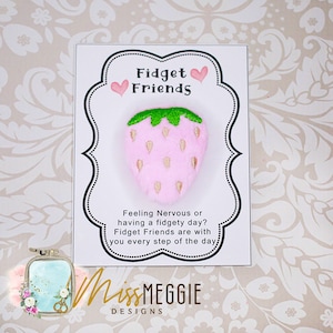 May include: A pink strawberry-shaped fidget toy with a green top, displayed on a card. The card reads "Fidget Friends" and includes text about managing fidgety days. The design includes a floral logo.
