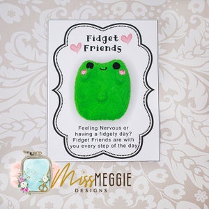 May include: A white card with the text "Fidget Friends" and a green frog plush toy. The card says "Feeling Nervous or having a fidgety day? Fidget Friends are with you every step of the day." The card also has the text "MissMEGGIE DESIGNS".