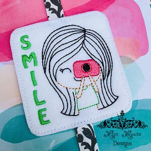 May include: White felt bookmark with green embroidered text "SMILE" and a black and white embroidered illustration of a girl with long hair holding a pink camera.