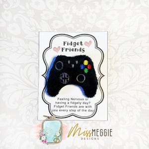 May include: A black plush controller with colourful buttons and the text "Fidget Friends" on a white card. The card also includes the text "Feeling Nervous or having a fidgety day? Fidget Friends are with you every step of the day."