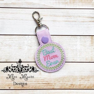 May include: Purple key chain with a silver key ring. The key chain has a round, embroidered patch with a green wreath and the text "Best Mom Ever" in pink, blue, and green lettering.