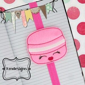 May include: Pink felt bookmark shaped like a macaron with a white stripe and a smiling face. The bookmark is attached to a pink elastic band.