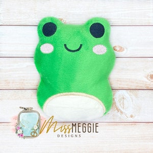 Puede incluir: Una rana de peluche verde con ojos negros, vientre blanco y una sonrisa cosida. La rana está sentada sobre una superficie de madera.