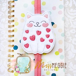Puede incluir: Un marcapáginas blanco con forma de gato con detalles de corazones rosas y una banda elástica rosa. El marcapáginas tiene detalles bordados, incluyendo una cara sonriente y mejillas rosas. El diseño está sobre un cuaderno con espiral.