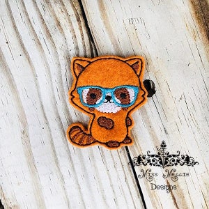 May include: An orange felt patch of a cartoon raccoon wearing blue spectacles. The patch has a white face, brown eyes, and a striped tail. The patch is on a wooden surface.