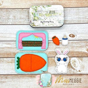 May include: A collection of felt bunny-themed items. Includes a tin with "My Bunny Home" text, a carrot, a bottle, a bunny, and two felt scenes: a stage and a large carrot. The items are in various colors, including pink, orange, and blue.