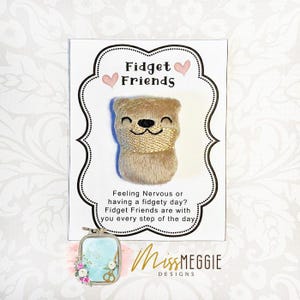 May include: A beige plush toy with a smiling face and a gold scarf is on a white card. The card says "Fidget Friends" and a message about managing fidgety days. The card is decorated with pink hearts and a decorative border.