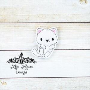 May include: White embroidered applique of a cartoon cat with pink ears. The cat is sitting with its legs tucked under its body. The applique is on a white wooden background.