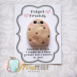 Puede incluir: Una tarjeta blanca con texto negro que dice "Fidget Friends" y "POSITIVE POTATO I maybe be a tiny potato but I believe in you!" Una patata de peluche marrón con una cara sonriente está unida a la tarjeta. La tarjeta tiene un borde negro y corazones rosas.