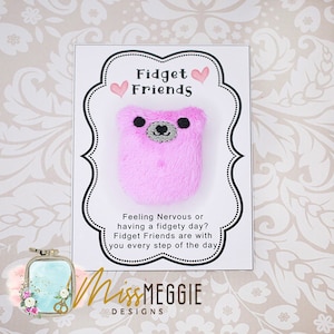 Può includere: Un giocattolo antistress a forma di orso rosa e peloso con occhi neri e un naso grigio. Il giocattolo è su una carta bianca con il testo "Fidget Friends" e "Feeling Nervous or having a fidgety day? Fidget Friends are with you every step of the day." La carta ha anche il testo "MissMEGGIE DESIGNS".