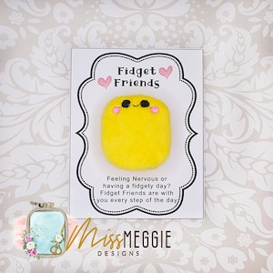 Peut inclure: Un jouet de stimulation en feutre jaune avec un visage souriant et des joues roses. Le jouet est sur une carte blanche avec le texte "Fidget Friends" et "Feeling Nervous or having a fidgety day? Fidget Friends are with you every step of the day." La carte porte également le texte "MissMEGGIE DESIGNS".
