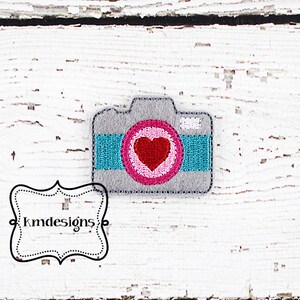 May include: A grey camera with a pink and white circle containing a red heart in the centre. The camera has a turquoise stripe across the bottom.