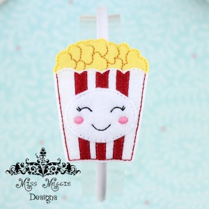 May include: A white headband featuring a felt popcorn bucket applique with red and white vertical stripes, yellow popcorn kernels, and a white oval patch with a stitched smiling face, pink cheeks, and closed eyes. The text Miss Meggie Designs is visible.