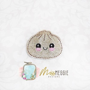 Könnte beinhalten: Ein glitzernder, goldener Stickaufnäher in Form eines Teigtäschchens mit lächelndem Gesicht, schwarzen Augen und rosa Wangen. Das Design hat einen gestickten Rand und befindet sich über einem kleinen Logo mit der Aufschrift Miss Meggie Designs.