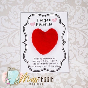 May include: A white card with black text that reads "Fidget Friends" and "Feeling Nervous or having a fidgety day? Fidget Friends are with you every step of the day". A red fuzzy heart is attached to the card.